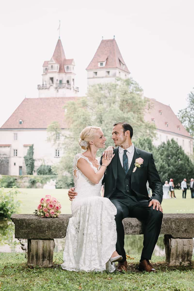 Ein Brautpaar sitzt auf einer Bank vor einem Schloss.