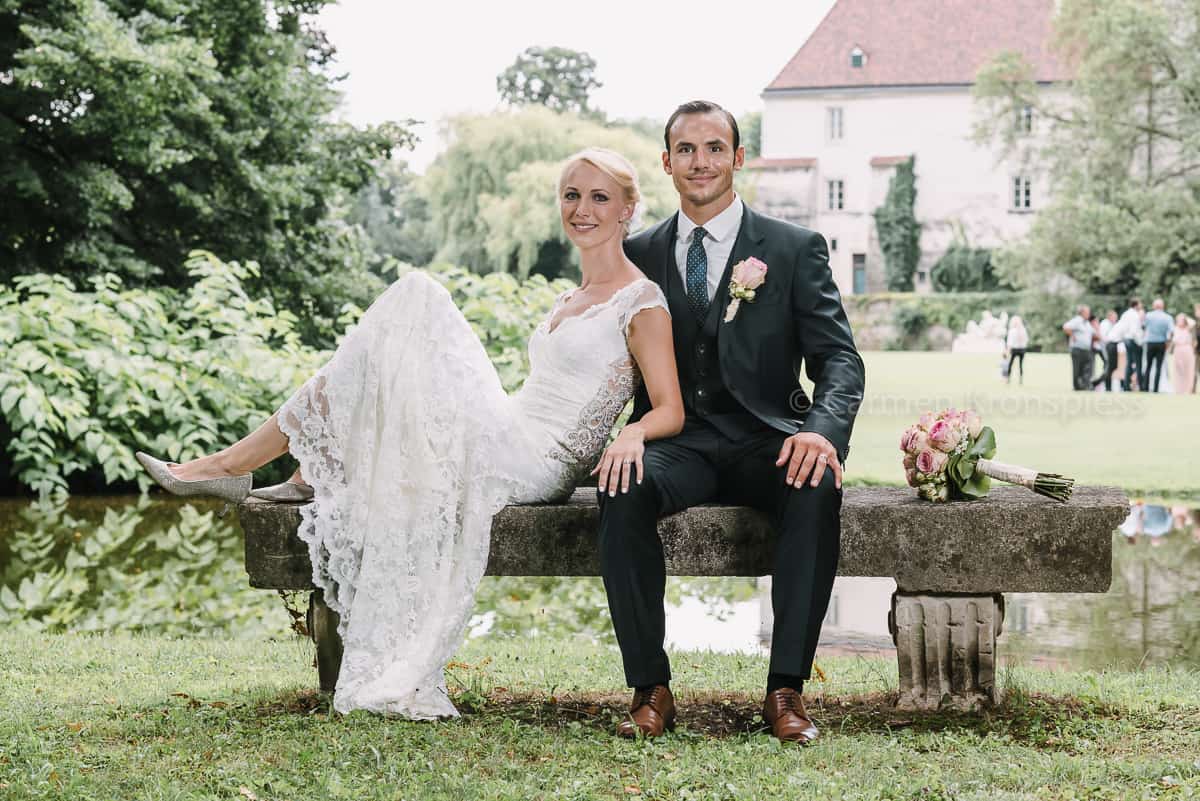 Ein Brautpaar sitzt auf einer Bank vor einem Teich.