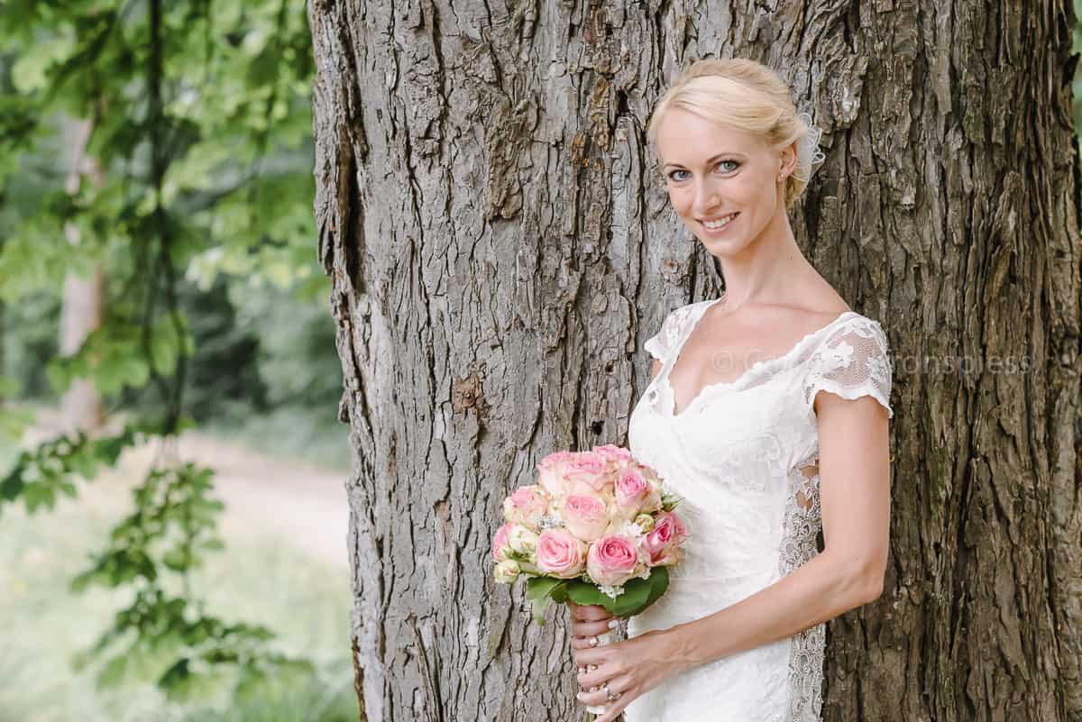 Eine Braut lehnt mit ihrem Blumenstrauß an einem Baum.