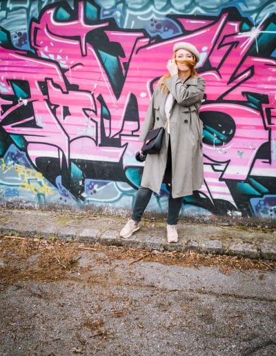 Eine Frau im Trenchcoat steht vor einer Graffitiwand.