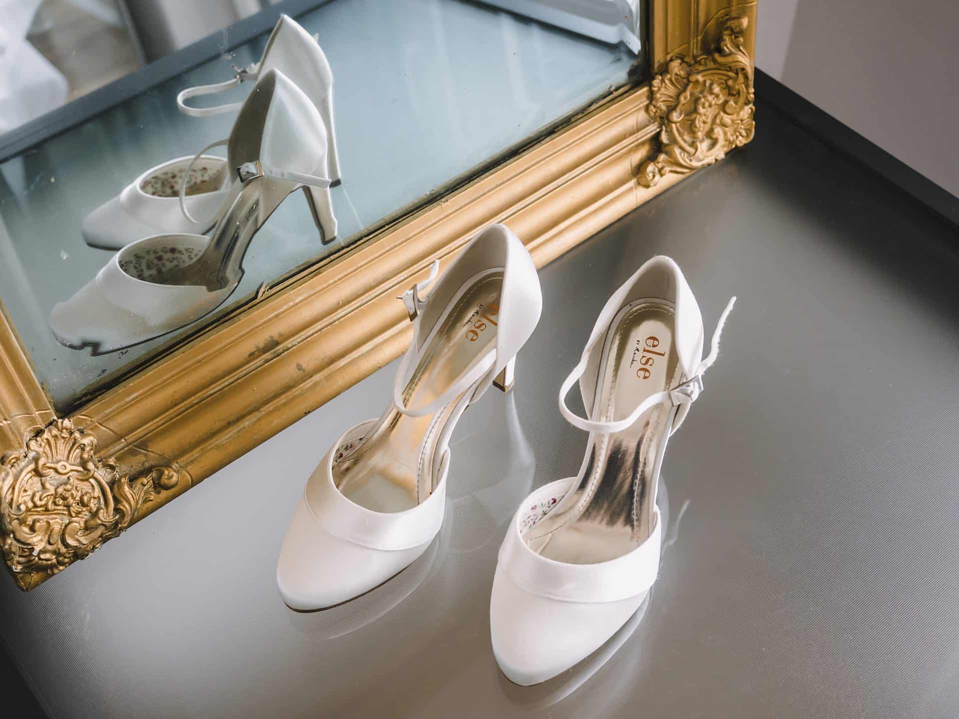 Ein Paar weiße Hochzeitsschuhe vor einem Spiegel.