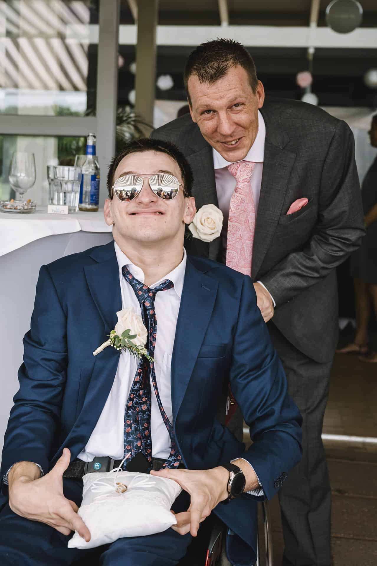 Ein Mann in Anzug und Sonnenbrille posiert mit einem Mann im Rollstuhl.