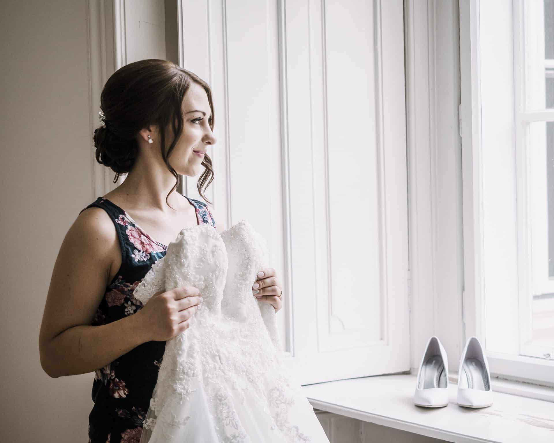Eine Braut schaut vor einem Fenster auf ihr Hochzeitskleid.
