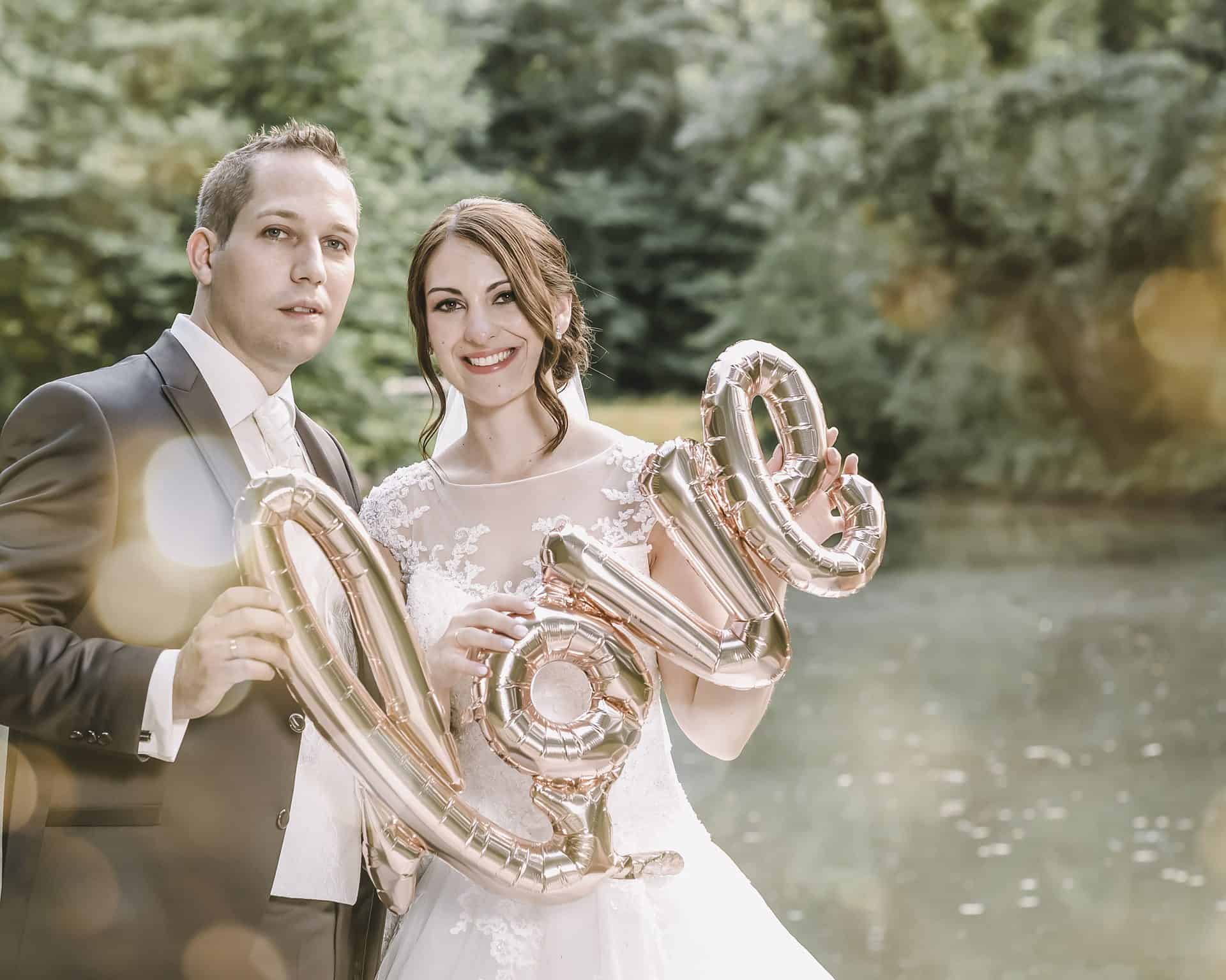 Eine Braut und ein Bräutigam halten Liebesballons vor einem Teich.