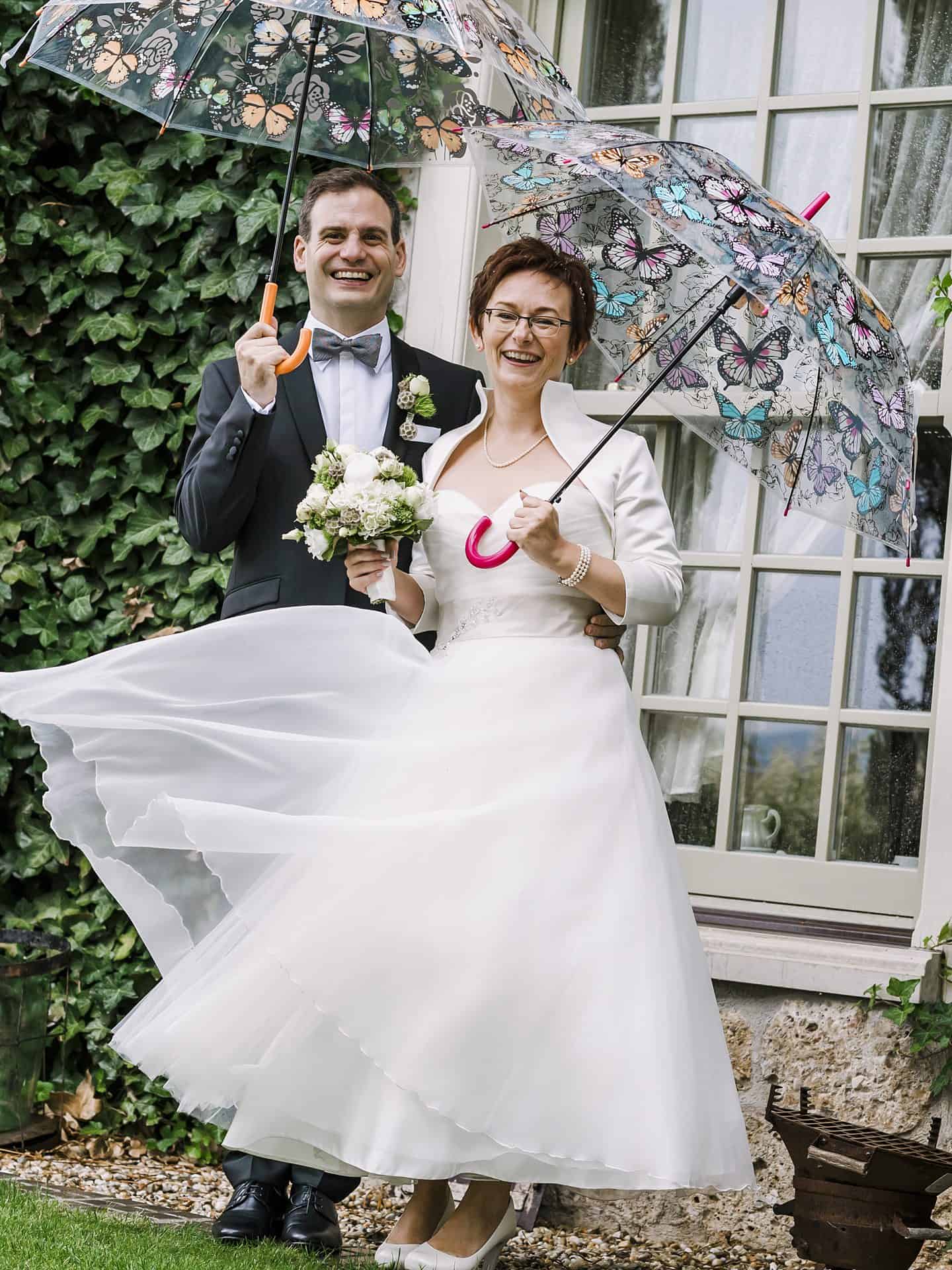 Eine Braut und ein Bräutigam halten Regenschirme vor einem Haus.