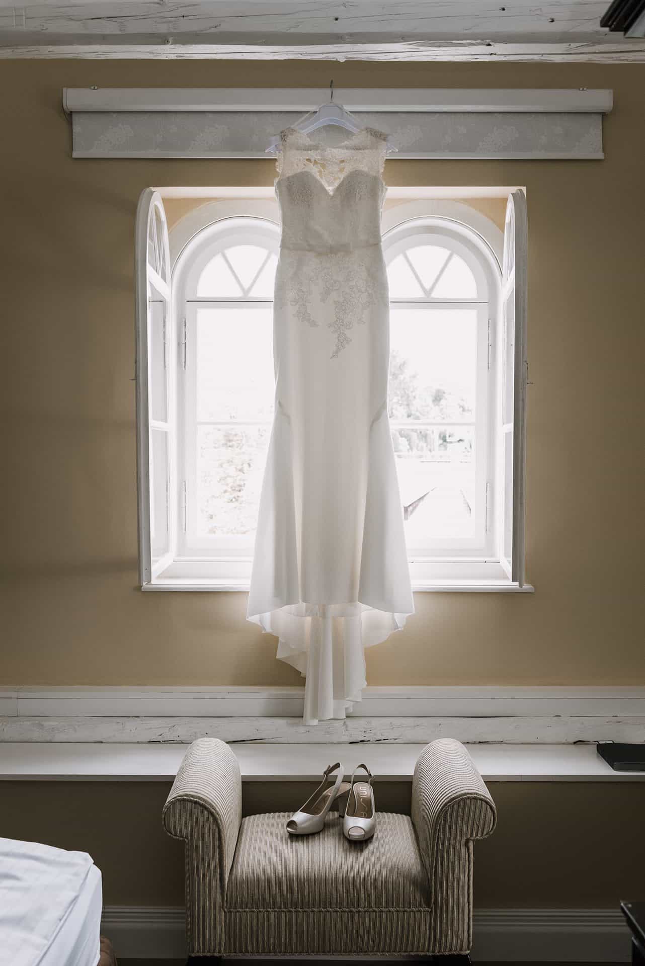 Ein Hochzeitskleid hängt vor einem Fenster in einem Zimmer.
