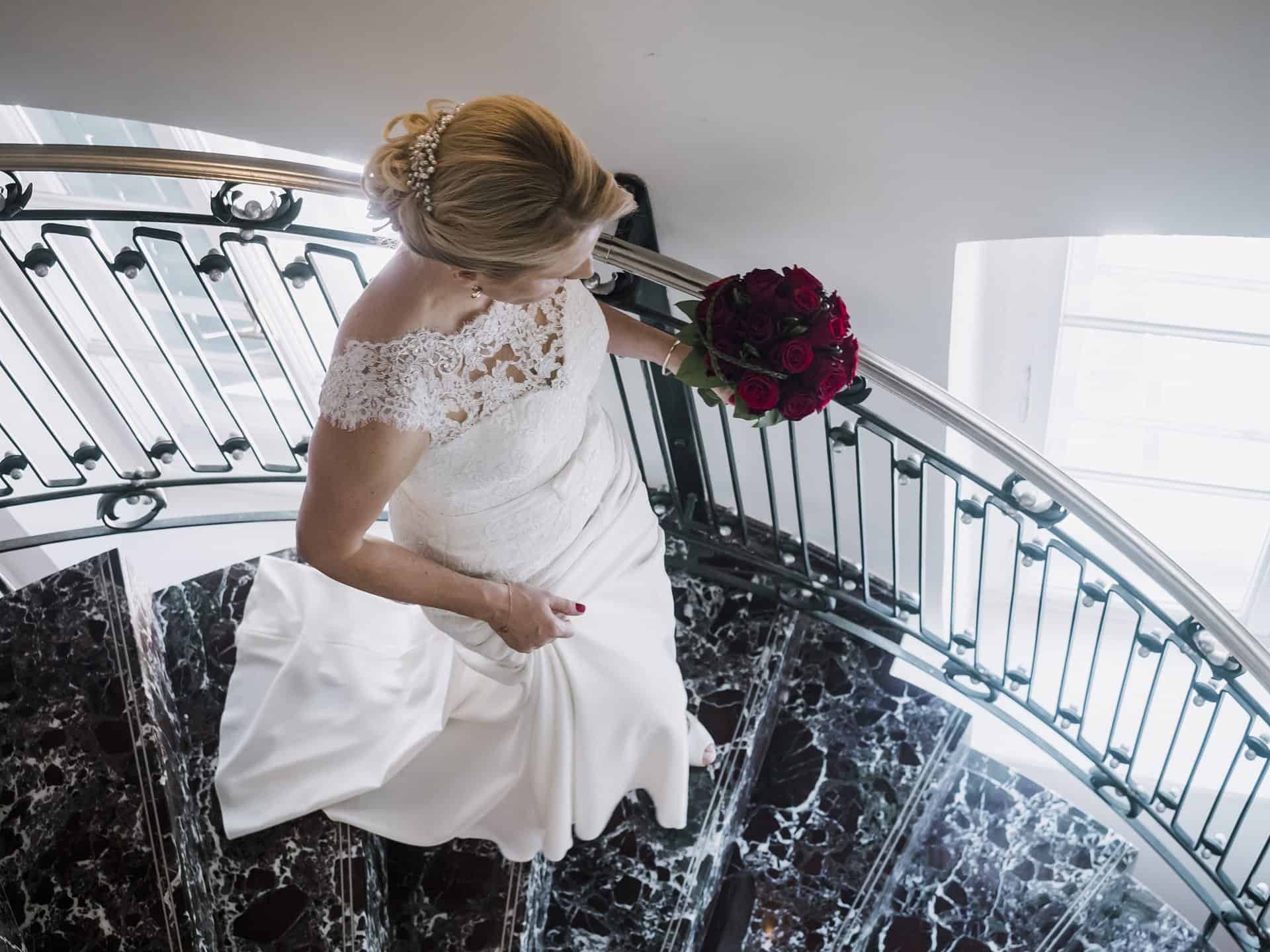 Eine Braut steht mit ihrem Blumenstrauß auf einer Wendeltreppe.