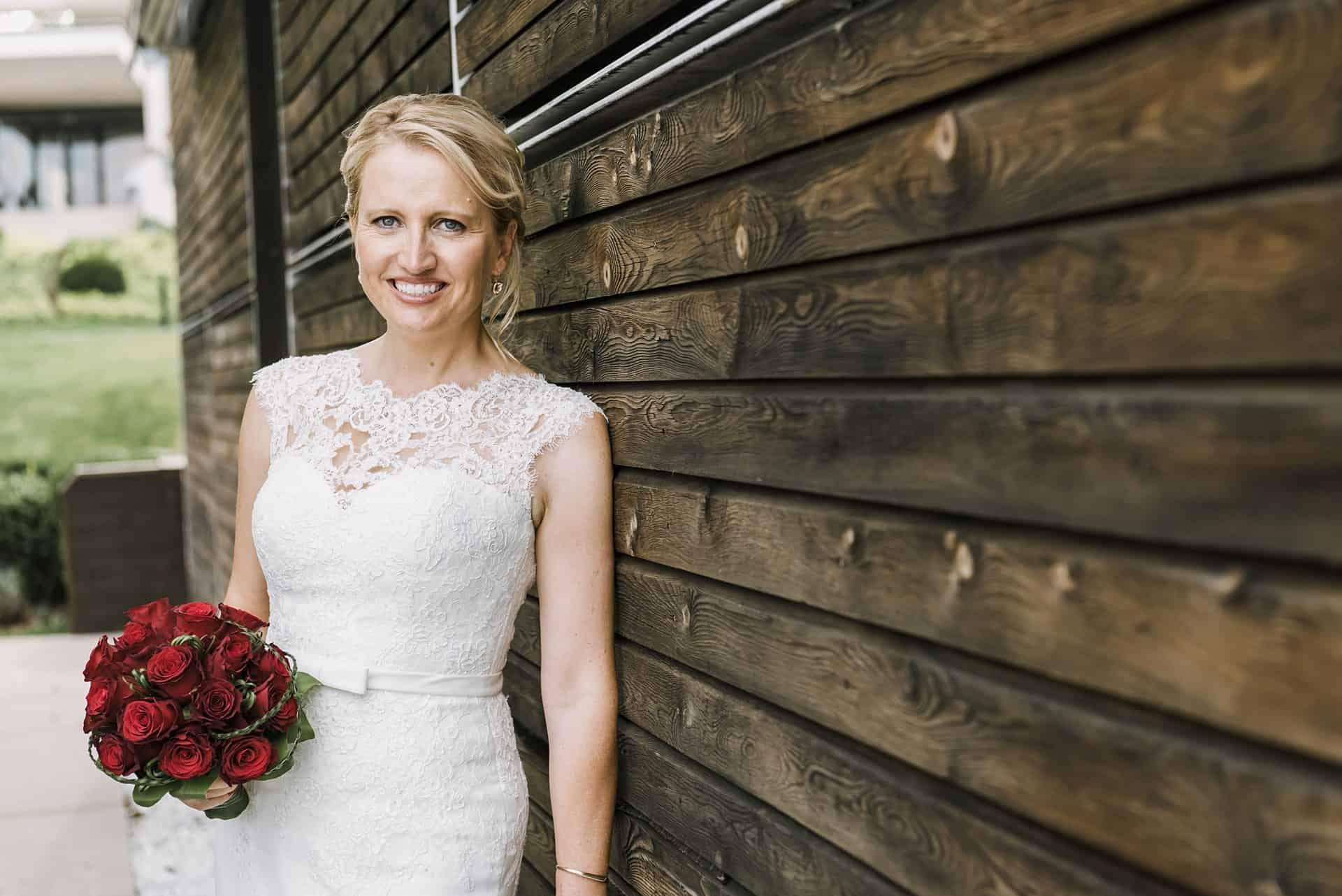 Eine Braut lehnt an einer Holzwand und hält einen Strauß roter Rosen in der Hand.