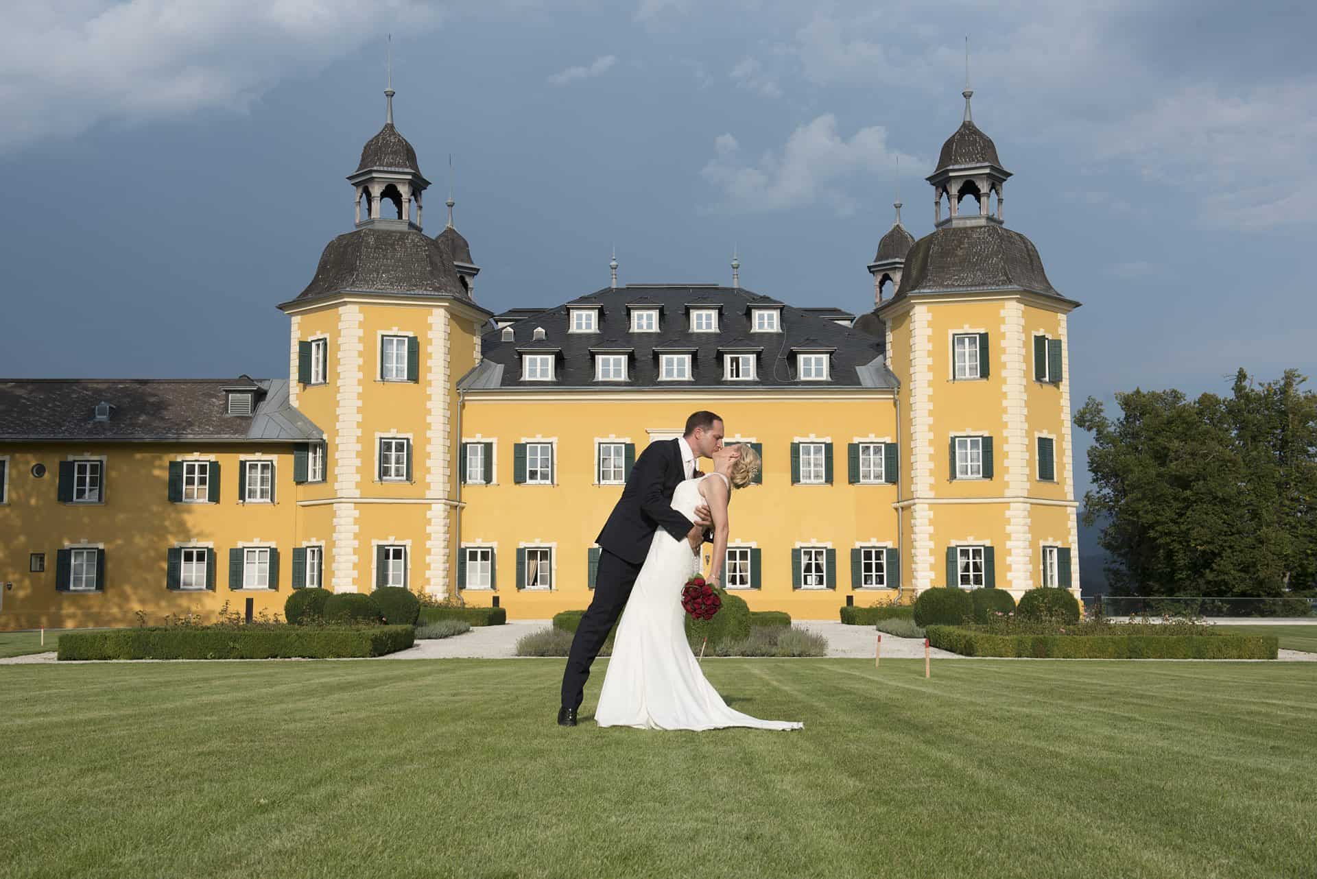 Ein Brautpaar küsst sich vor einem gelben Schloss.