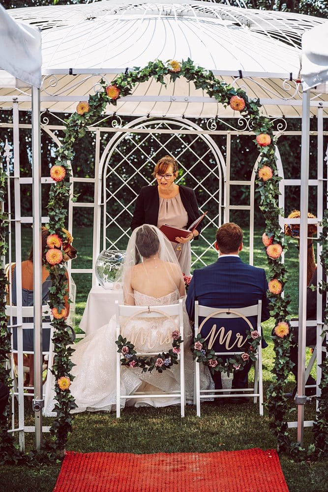 Eine Hochzeitszeremonie unter einem Pavillon mit Braut und Bräutigam, die auf Stühlen sitzen.