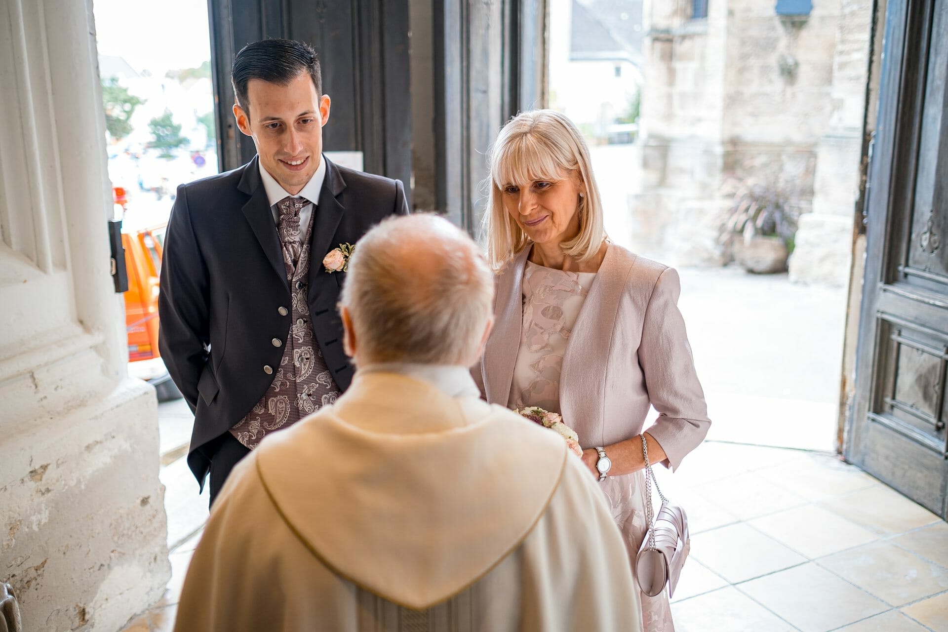 Ein Mann im Anzug und eine Frau im Kleid im Gespräch mit einem Priester.