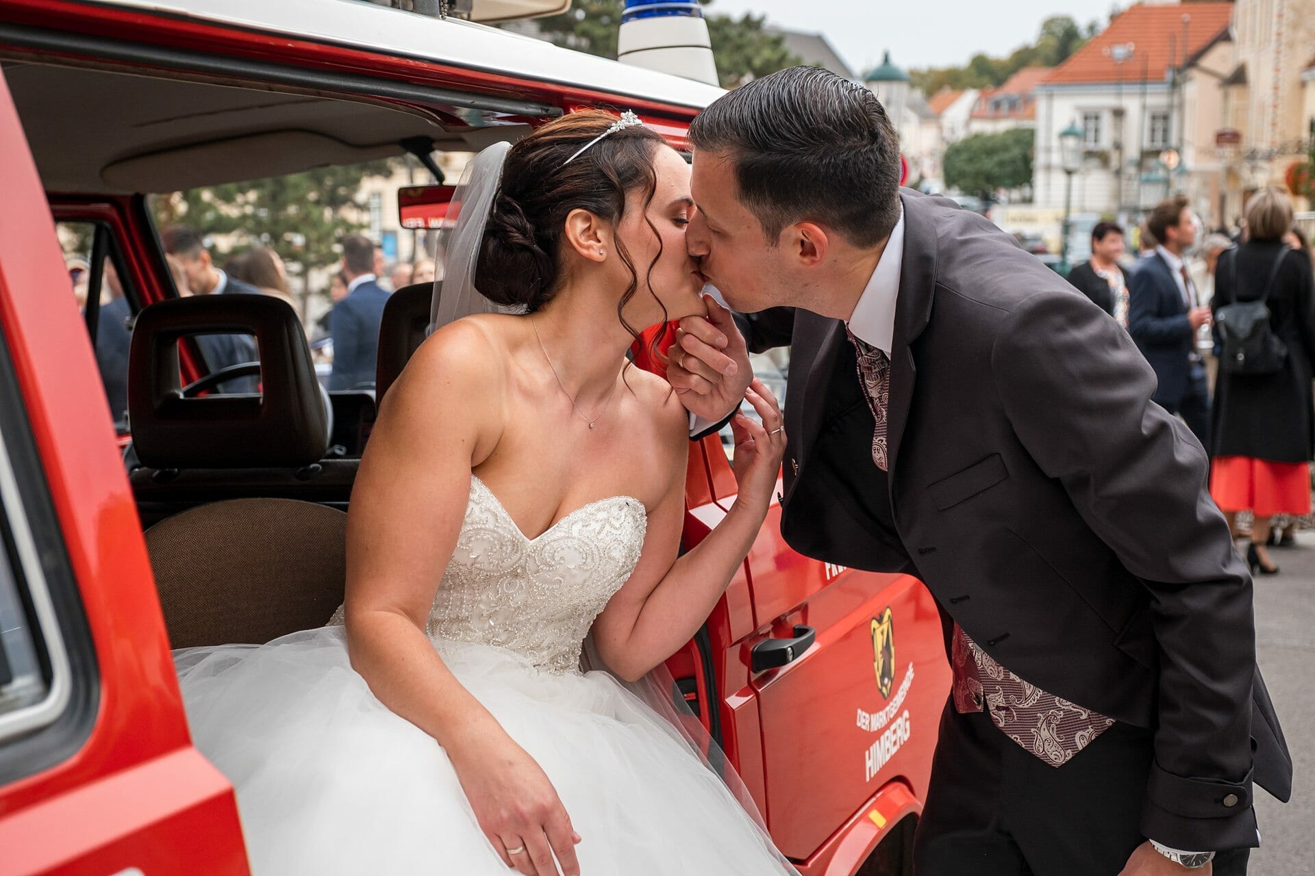 Eine Braut und ein Bräutigam küssen sich vor einem roten Auto.