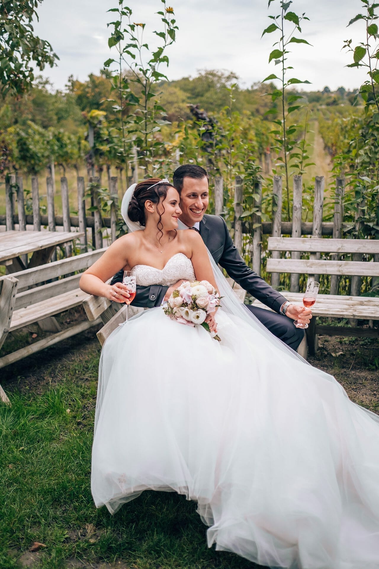 Eine Braut und ein Bräutigam sitzen auf einer Bank in einem Weinberg.