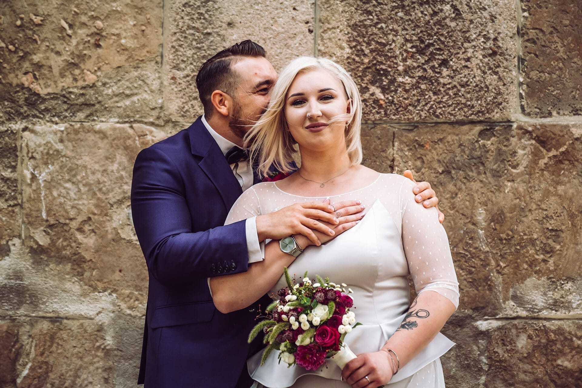 Eine Braut und ein Bräutigam umarmen sich vor einer Steinmauer.
