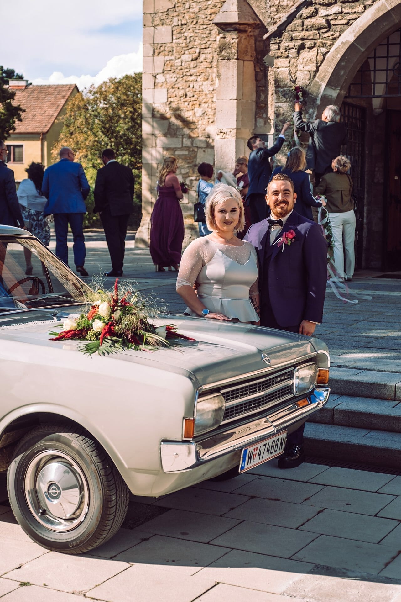 Ein Brautpaar steht neben einem alten Auto vor einer Kirche.